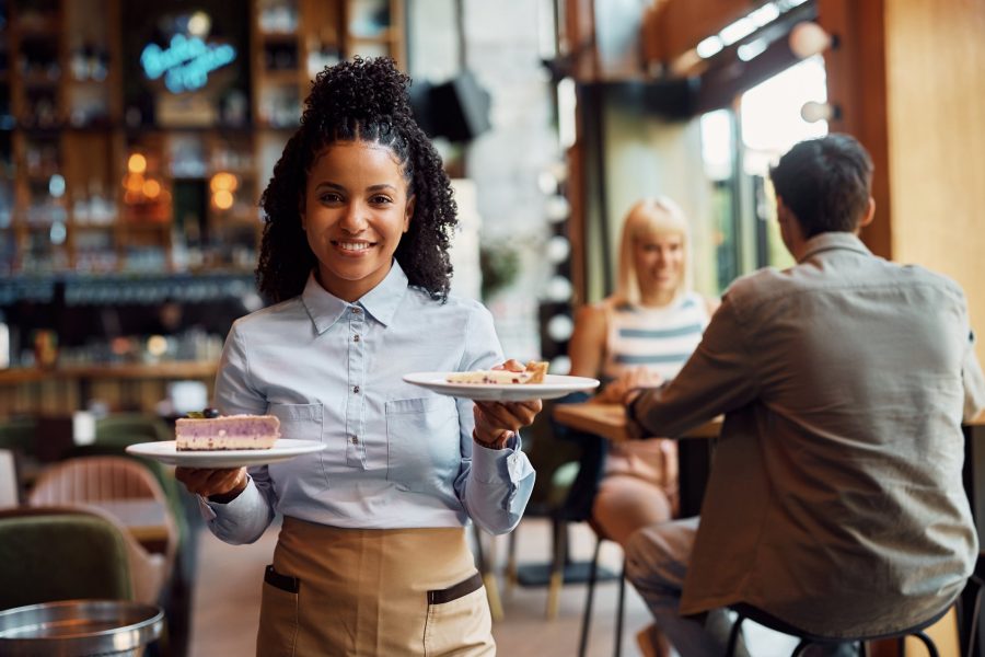 Happy black waitress serving dessert in a cafe and looking at camera. Happy African American waitress serving a cake while working in cafe and looking at camera.